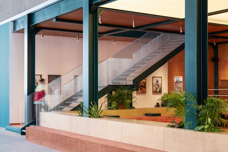 Metal mesh staircase ascending behind planted concrete planters with perforated orange acoustic panels beyond