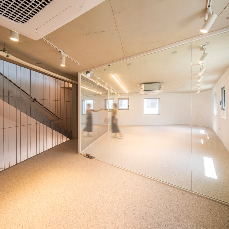 Interior space with glass partition, exposed plywood ceiling, track lighting, and blurred figures moving near the stairwell