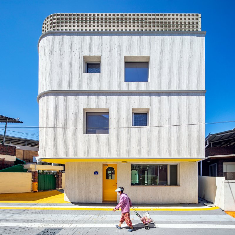 Street facade with vertical ribbed white cladding, square windows, and yellow ground floor with a person passing by