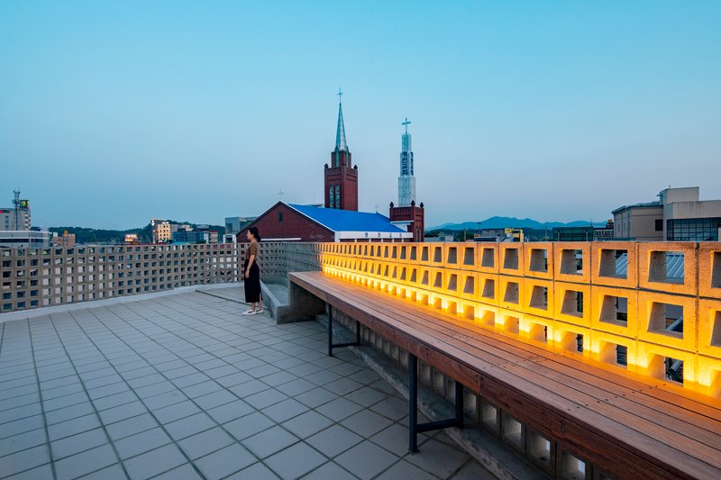 Rooftop terrace with timber bench and backlit perforated parapet facing church spires at twilight