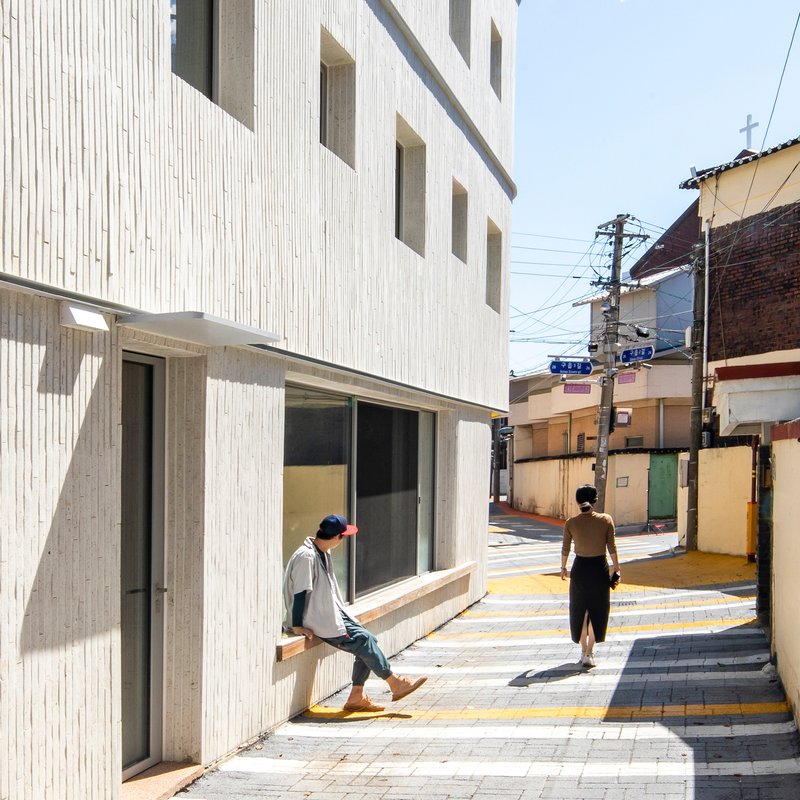 Entrance elevation with vertical cladding and yellow-striped pavement where two figures walk in morning light
