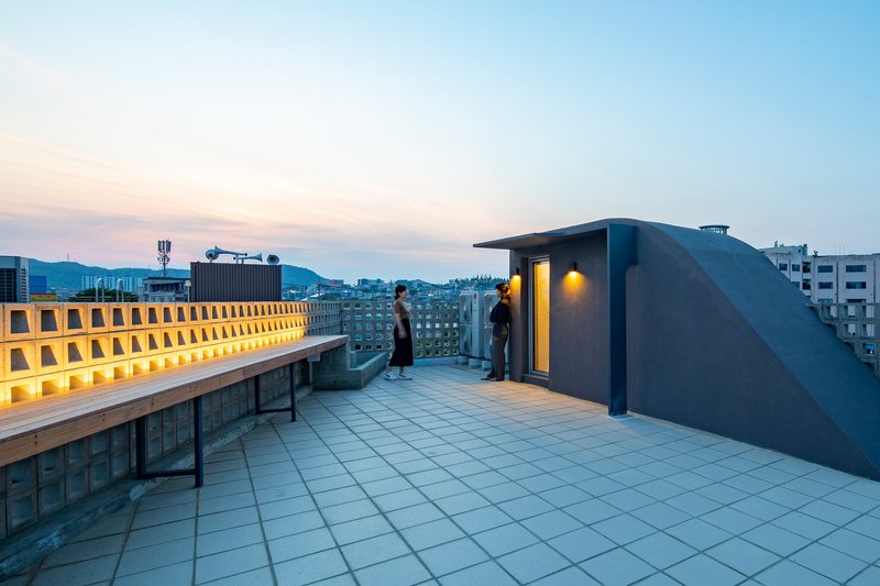 Rooftop terrace with tiled paving and illuminated perforated screen wall overlooking the neighborhood at dusk