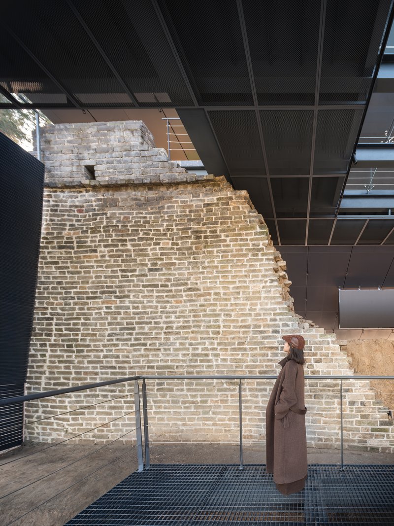 Visitor standing on walkway alongside preserved brick wall with visible deterioration and openings