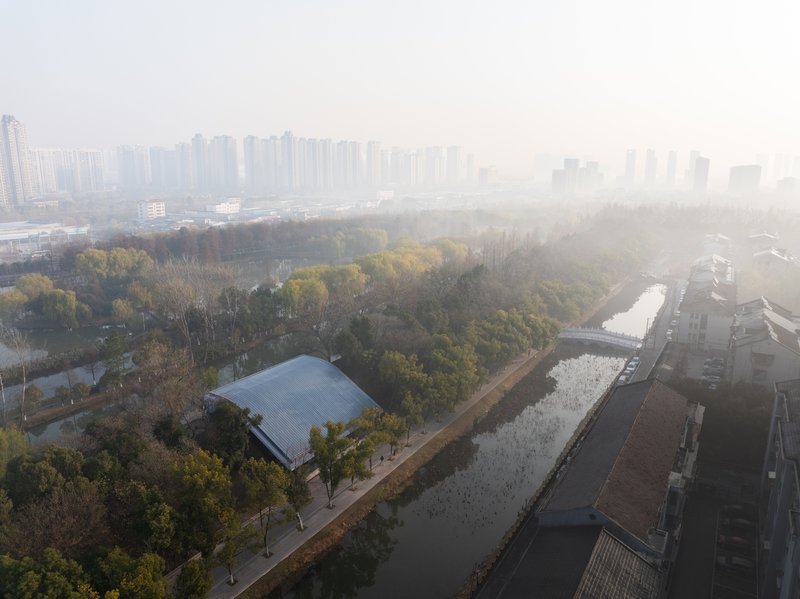 Aerial view of the arched metal roof nestled among autumn trees beside a canal and distant towers in heavy fog