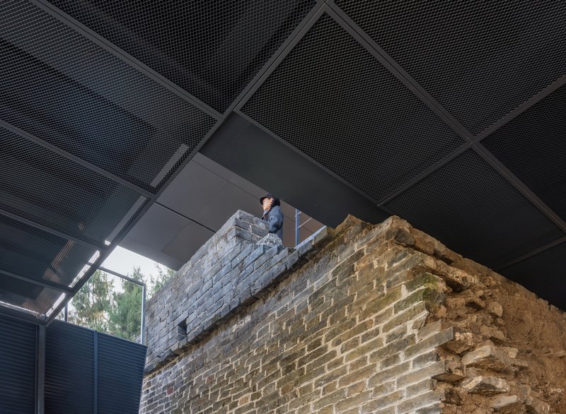 Visitor standing atop preserved brick wall remnant under perforated metal ceiling panels