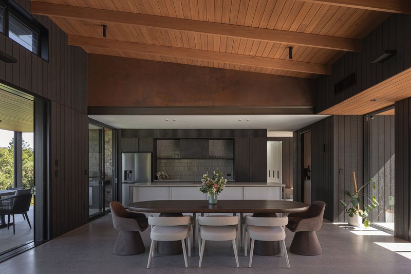 Open dining and kitchen area with timber ceiling beams and dark vertical wall cladding in afternoon light