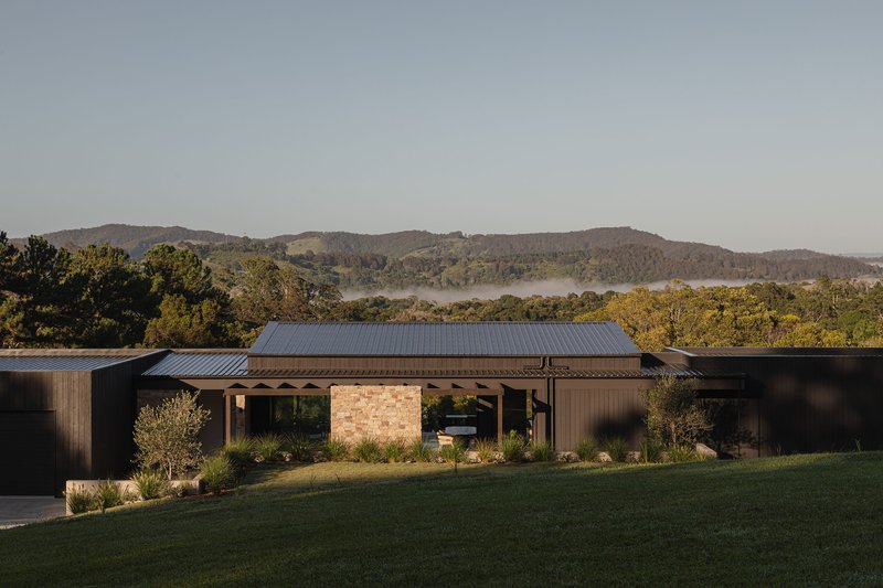 Front elevation with stone masonry core and vertical timber cladding set against forested hills in morning light