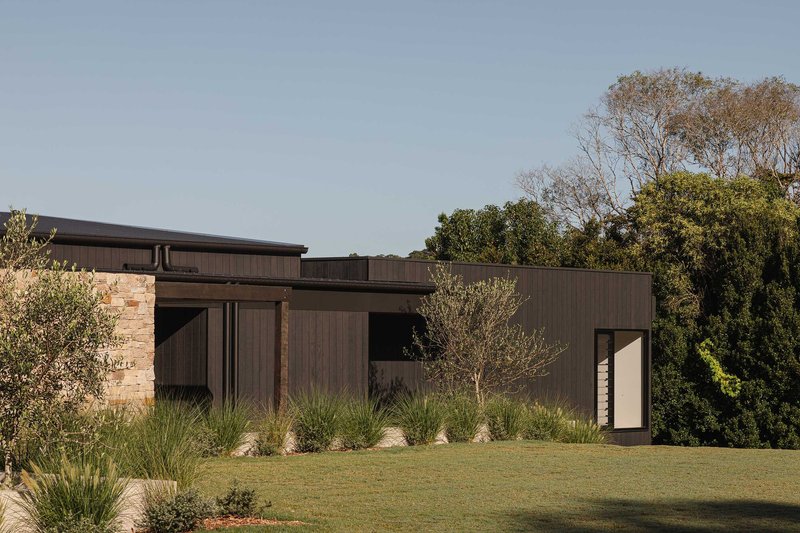 Side facade showing timber-clad volumes and glazed entry framed by native grasses and lawn