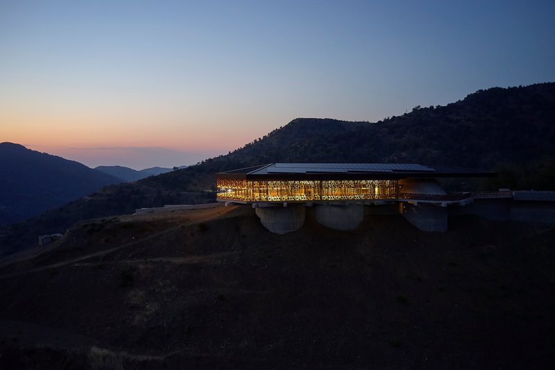 Illuminated glass facade perched on concrete piers against forested hills at dusk