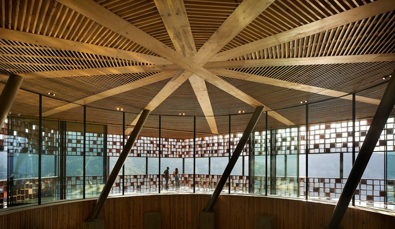 Interior view of radial timber beams converging at the ceiling center above glass-walled galleries with visitors