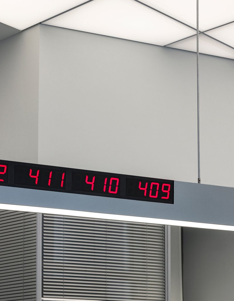 Red digital number display mounted on gray ceiling soffit above horizontal louvers