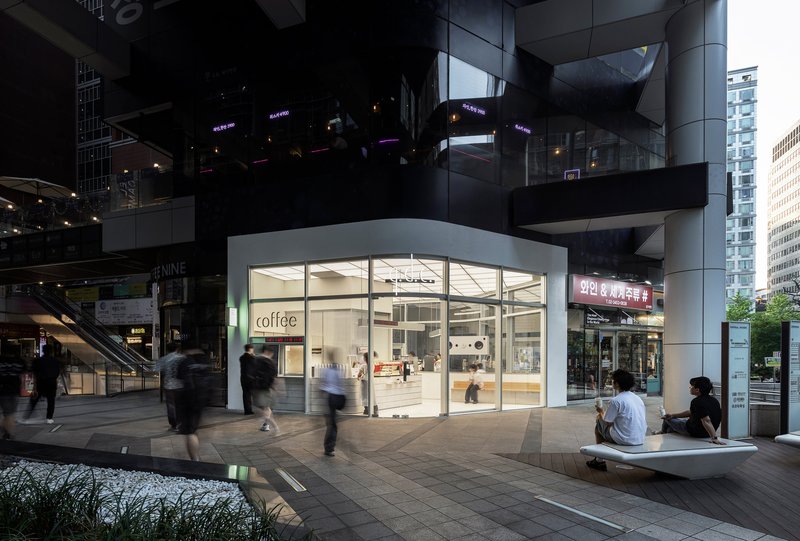 Glass storefront pavilion beneath a raised plaza structure at dusk with pedestrians passing by