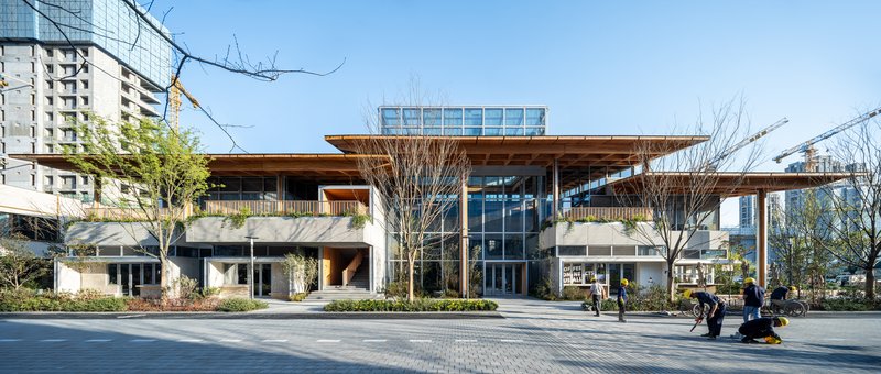 Street elevation showing timber roof overhang and central glass atrium with pedestrians in plaza