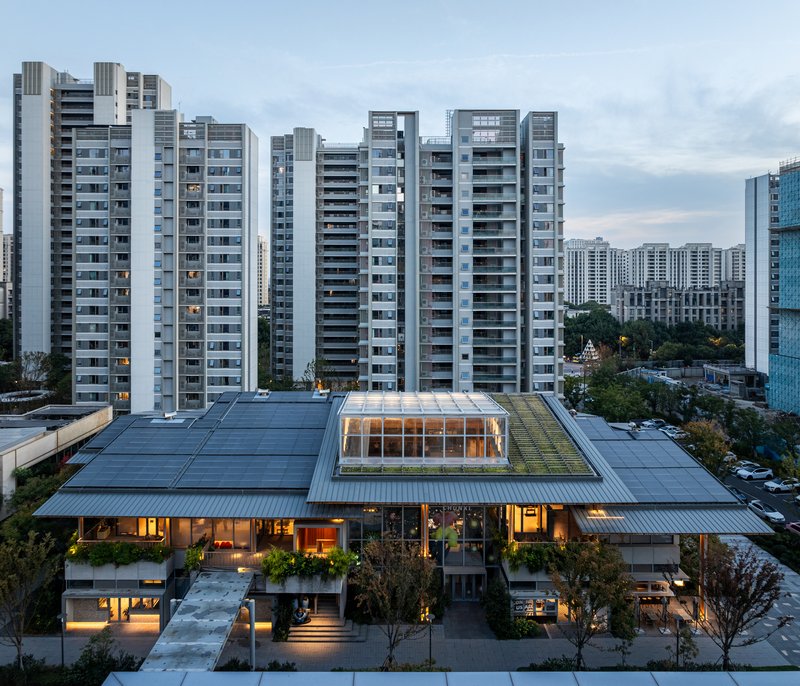 Aerial view of low-rise timber structure with roof terrace surrounded by residential towers at dusk