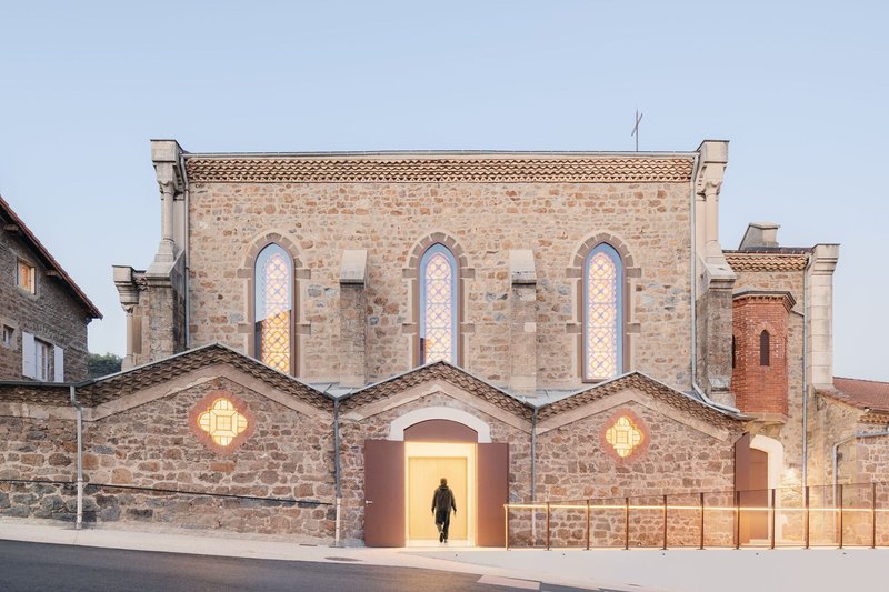 Front facade of stone church with arched entry, illuminated doorway and figure walking inside at dusk