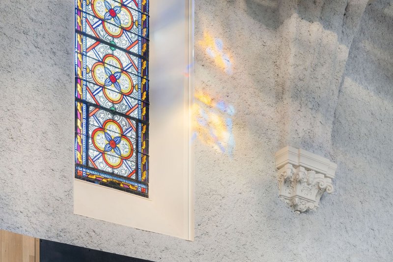 Stained glass window casting colored light onto a bare stone wall beside a carved capital