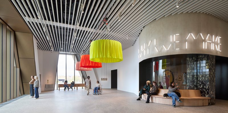 Double-height foyer with yellow and orange pendant lights and visitors beneath curved slatted ceiling