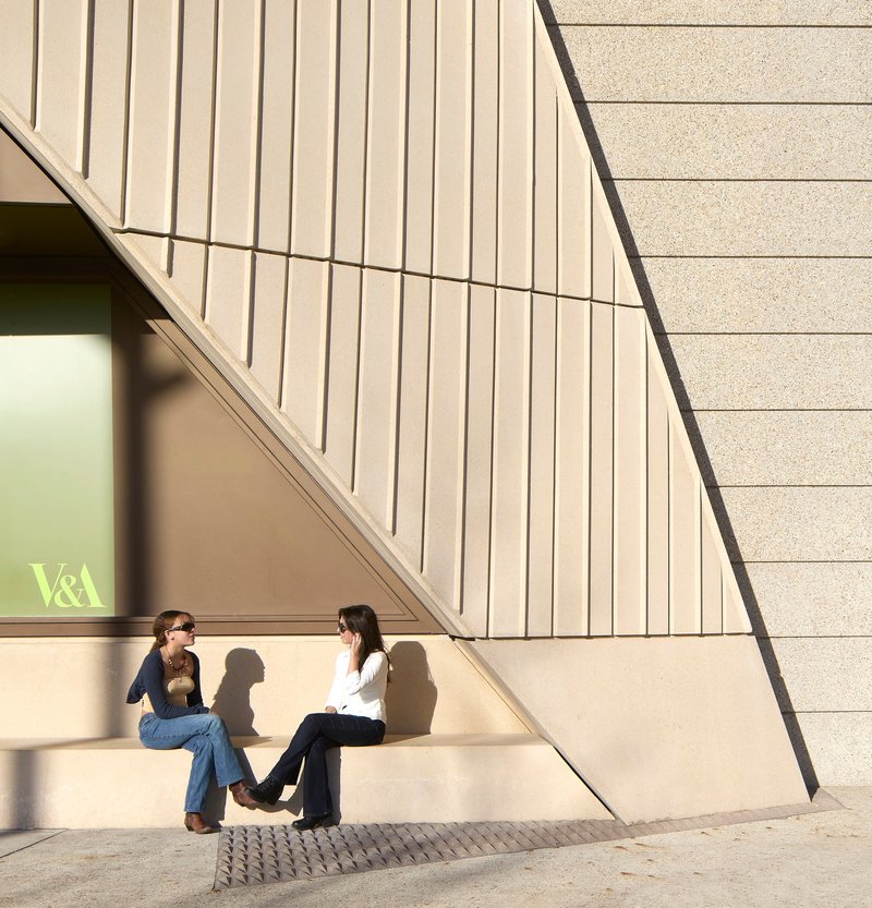 Exterior corner with vertical fluted panels and textured stone where two visitors sit in midday sun