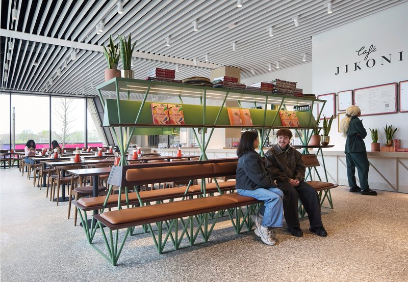 Cafe seating with green metal bench and shelving unit beneath a slatted ceiling with visitors nearby