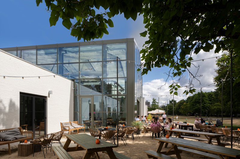 Two-story glass box addition connecting to white masonry volume with outdoor terrace seating under tree canopy