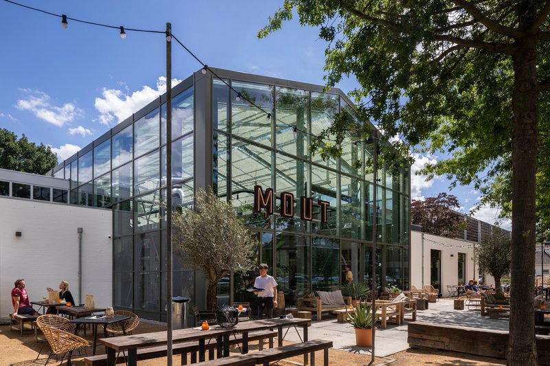 Glass-walled brewery pavilion with outdoor terrace seating under a blue sky