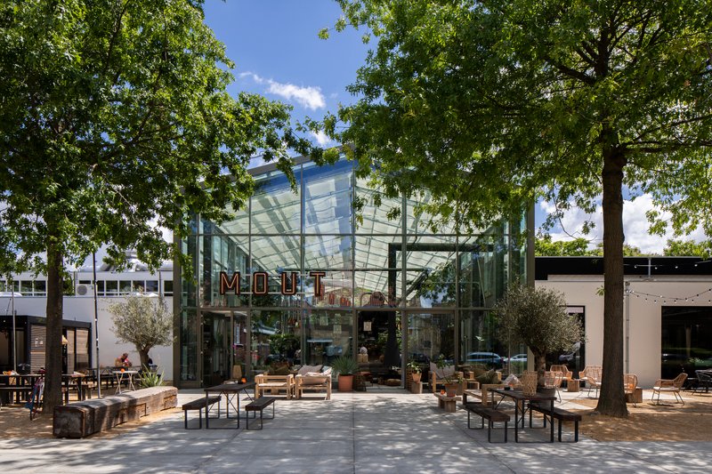 Glass pavilion entrance with steel frame and translucent canopy roof beneath mature trees on a sunny day