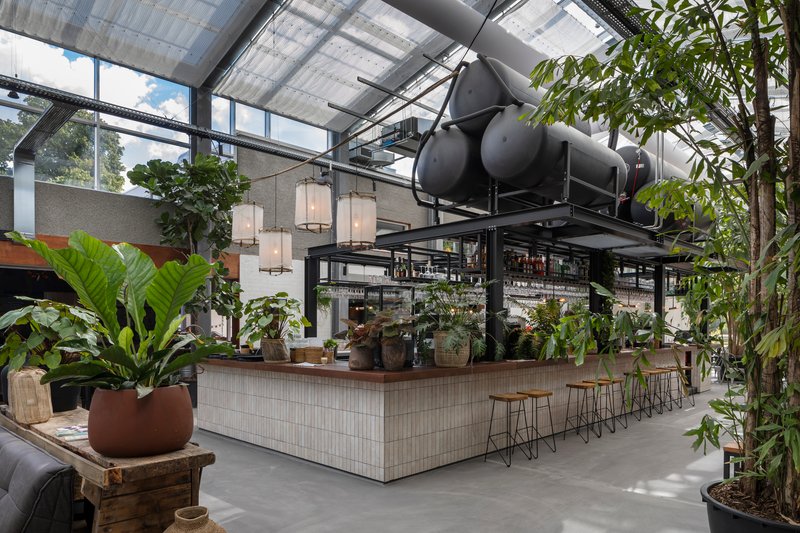 Tiled bar counter beneath exposed mechanical ductwork and skylights with potted plants throughout