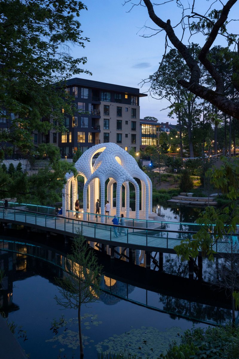 Illuminated pavilion reflected in still pond water at dusk with residential buildings beyond