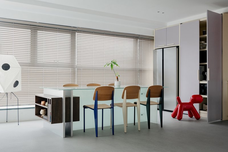 Dining area with frosted glass table, mismatched chairs, and horizontal blinds filtering daylight from floor-to-ceiling windows