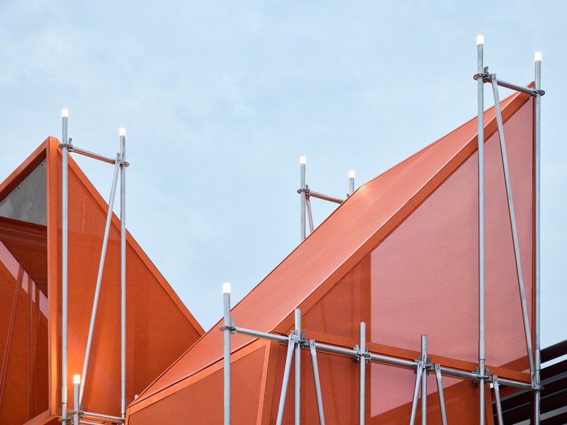 Angled orange metal canopy panels supported by scaffolding towers against a pale sky