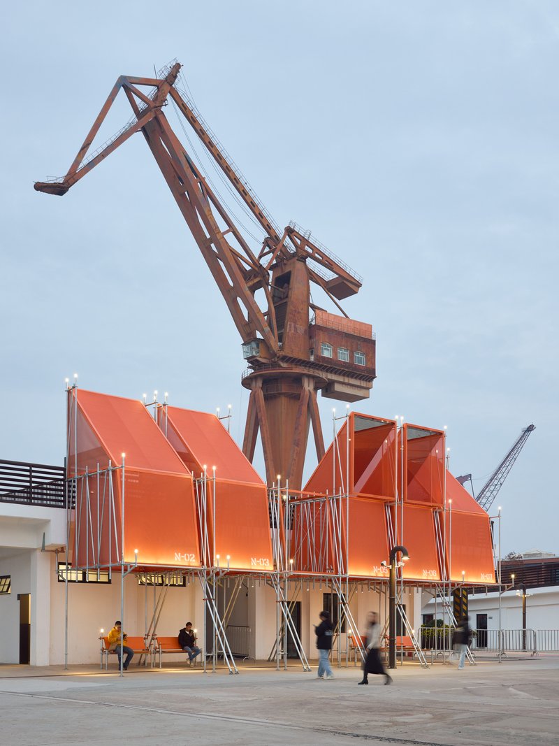 Orange metal canopy structures beneath a historic shipyard crane as visitors walk through the plaza