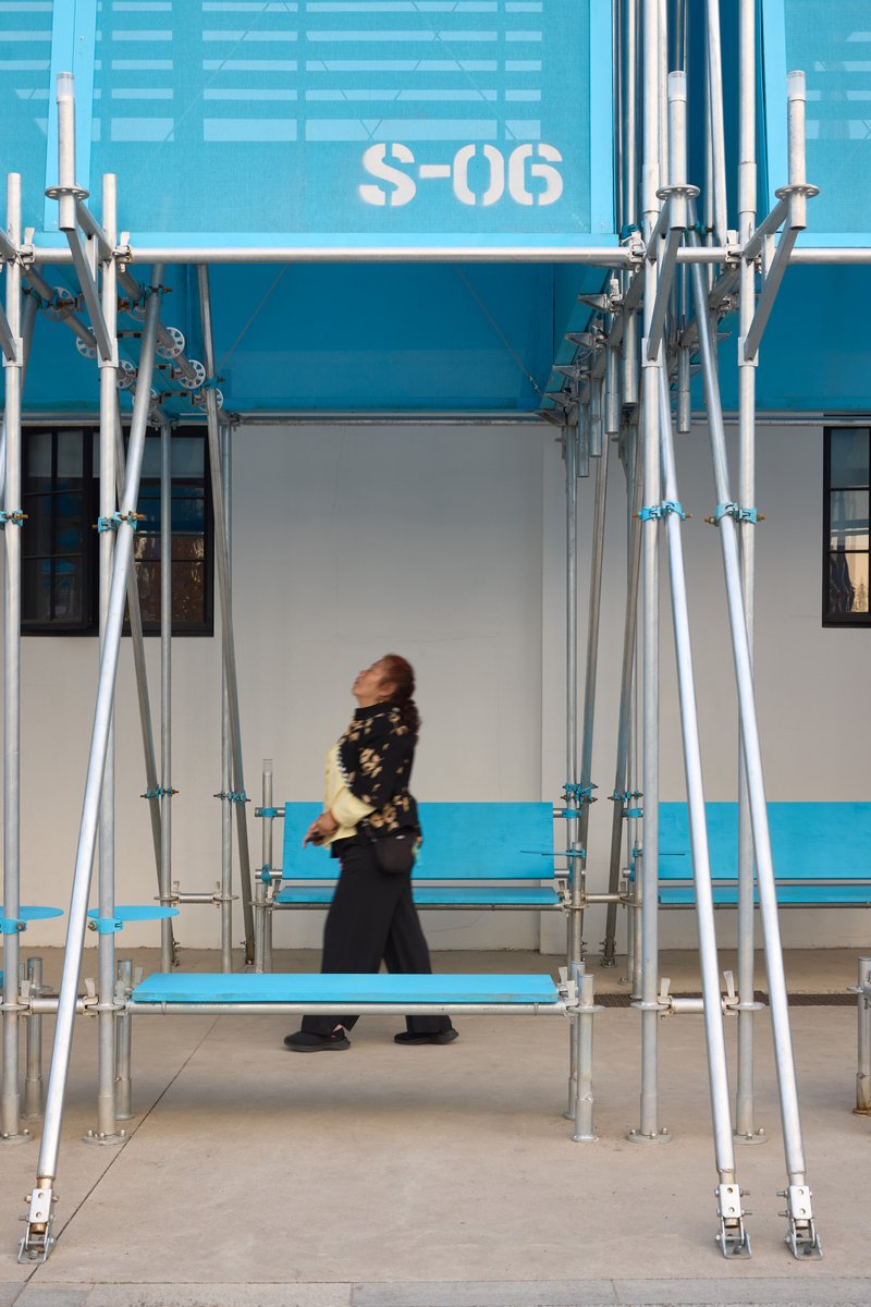 Woman walking past scaffolding columns beneath blue modular unit labeled S-06