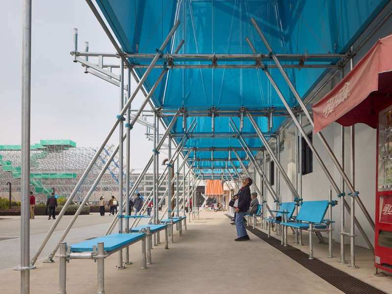 Covered walkway with scaffolding framework, blue canopy overhead, and benches where visitors pause in daylight