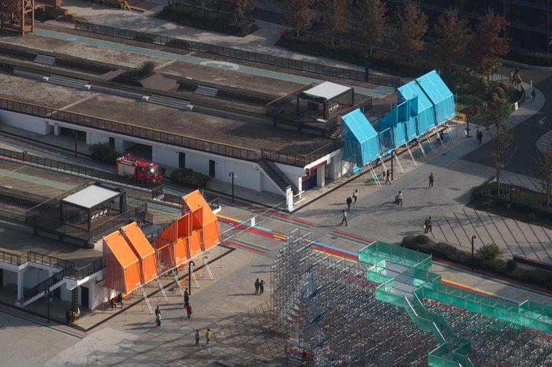Aerial view of three colored pavilions on a plaza with pedestrians and nearby rail tracks