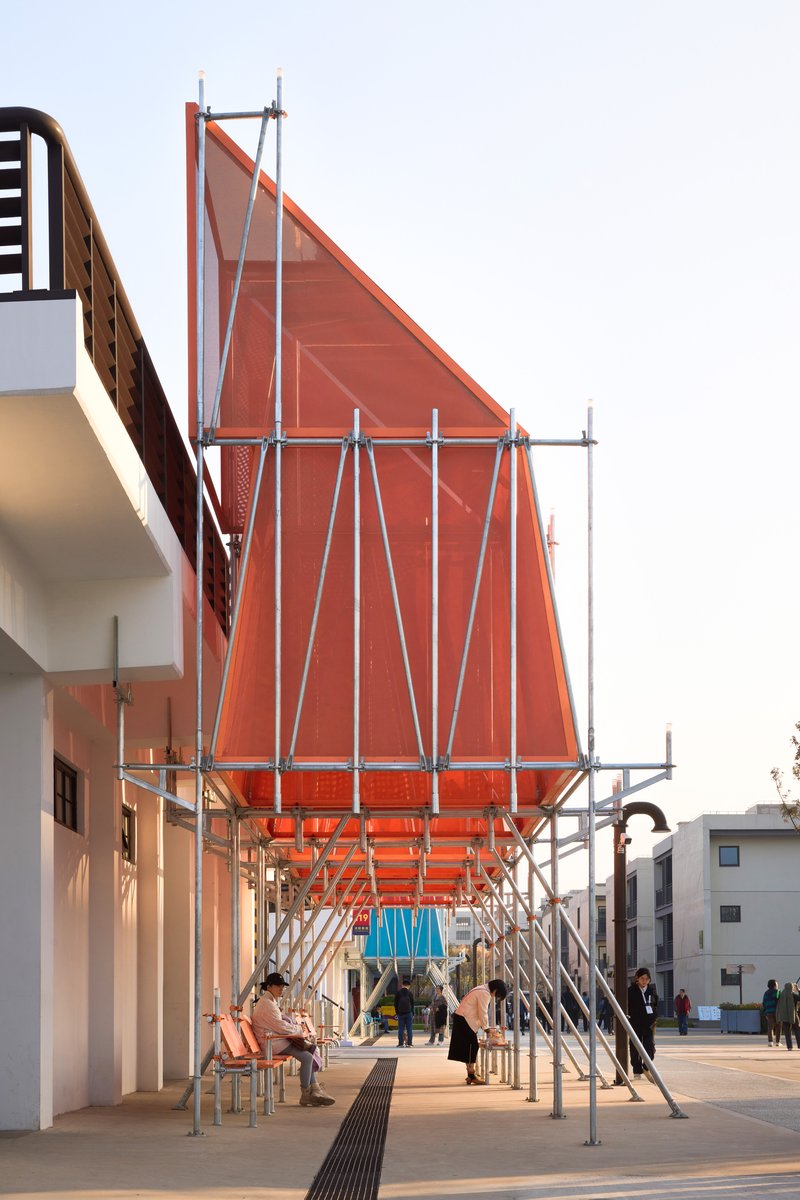Underside of orange translucent canopy supported by tubular steel scaffolding with people walking below