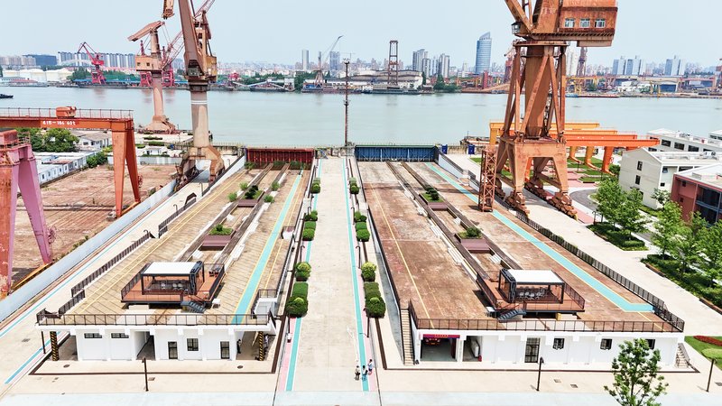 Elevated view of linear industrial site with warehouses, planted rows, and harbor cranes beyond
