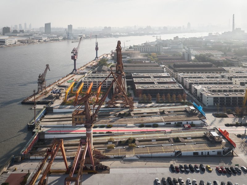 Elevated view of orange scaffold structures and a blue pavilion amid industrial harbor cranes