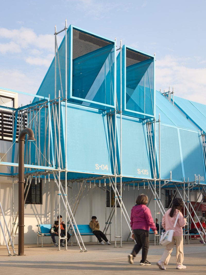 Blue polycarbonate pavilion with triangular gables and scaffolding framework as pedestrians walk past