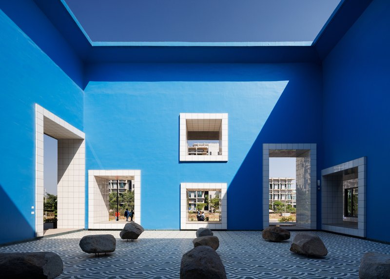 Blue-painted courtyard with white-framed windows, scattered rocks on tiled paving, and views through to planted greenery