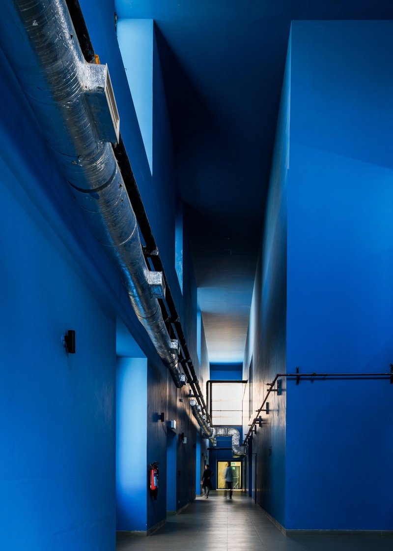 Blue-painted service corridor with exposed ductwork and a figure walking toward the lit end