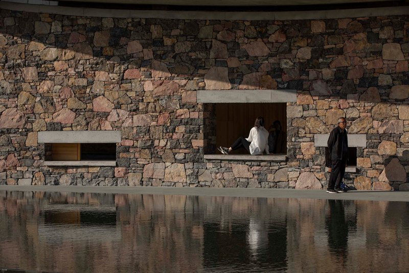 Fieldstone facade with recessed concrete-framed openings reflected in the still surface of a pool