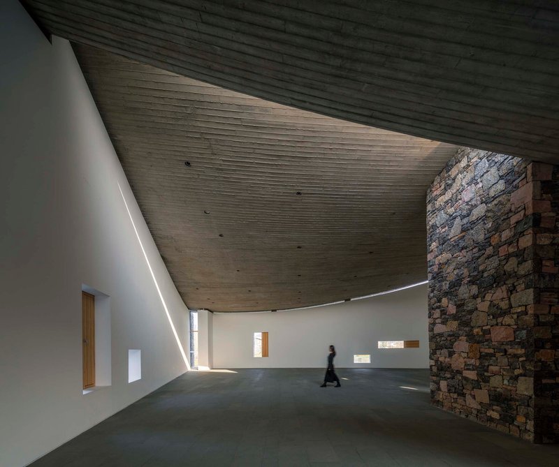 Interior hall with sloping timber ceiling and stone wall as a figure walks through sunlight