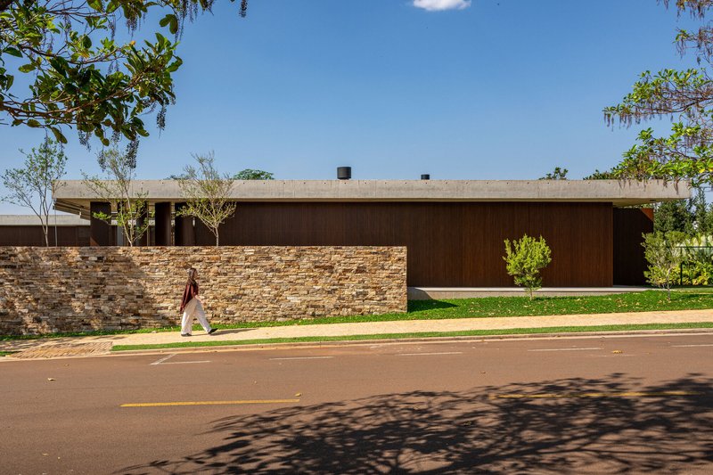 Street view of layered stone wall and timber-clad volume with a pedestrian walking past under tree shade