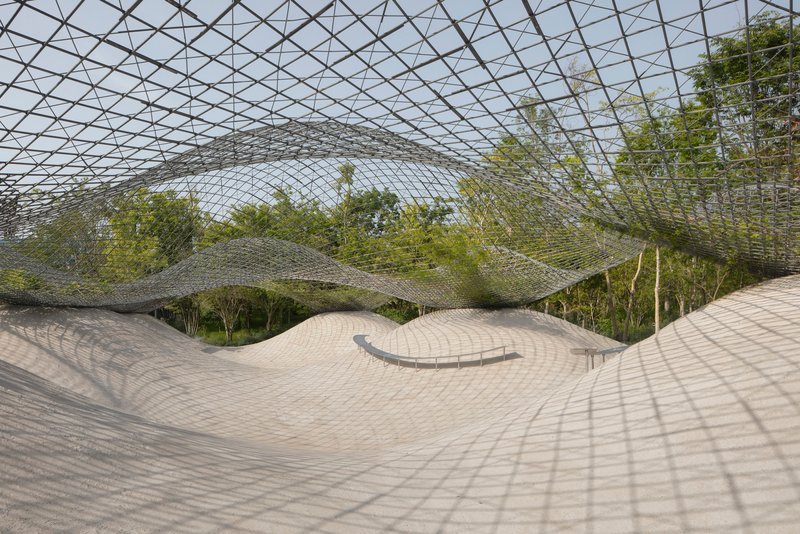 Undulating paved courtyard beneath a diagrid metal canopy with dappled shadows from overhead trees