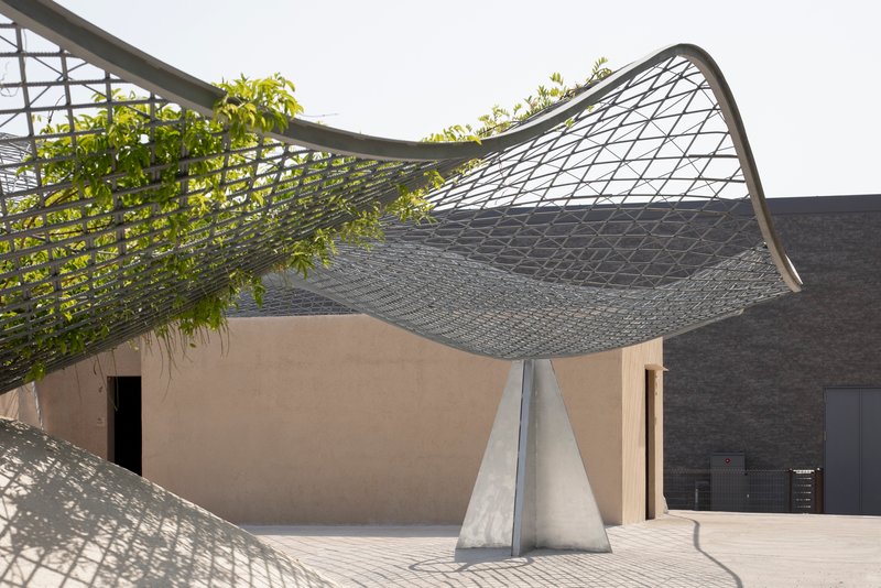 Curved metal mesh canopy rising from concrete supports over a courtyard with planted greenery