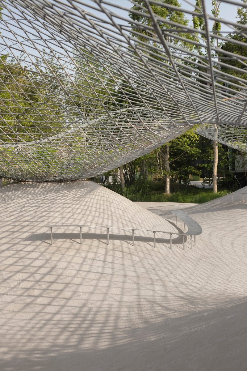 Metal mesh roof extending over paved plaza with young trees casting geometric shadows below