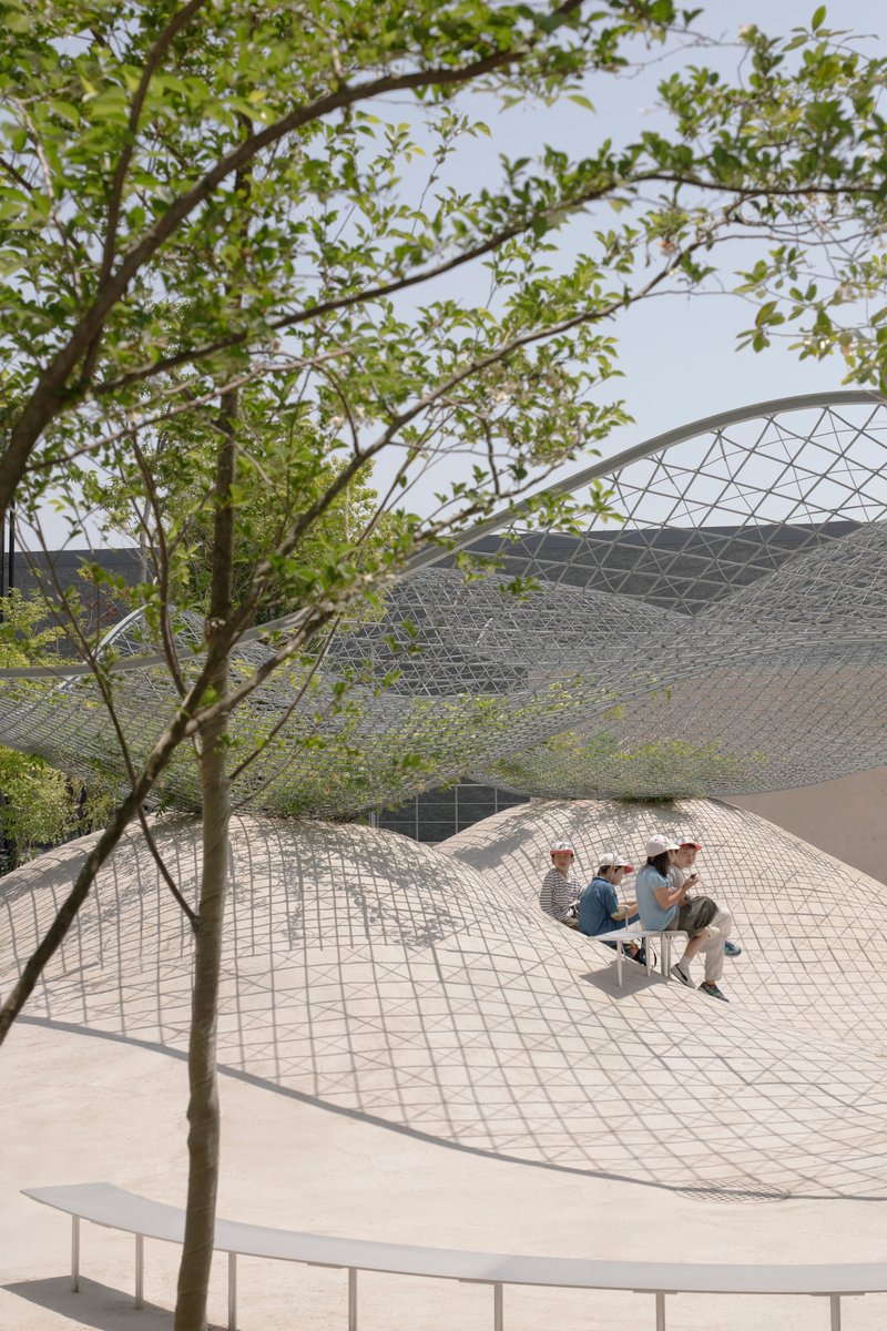 Two visitors seated in a sunken tiled courtyard framed by spring foliage and metal canopy