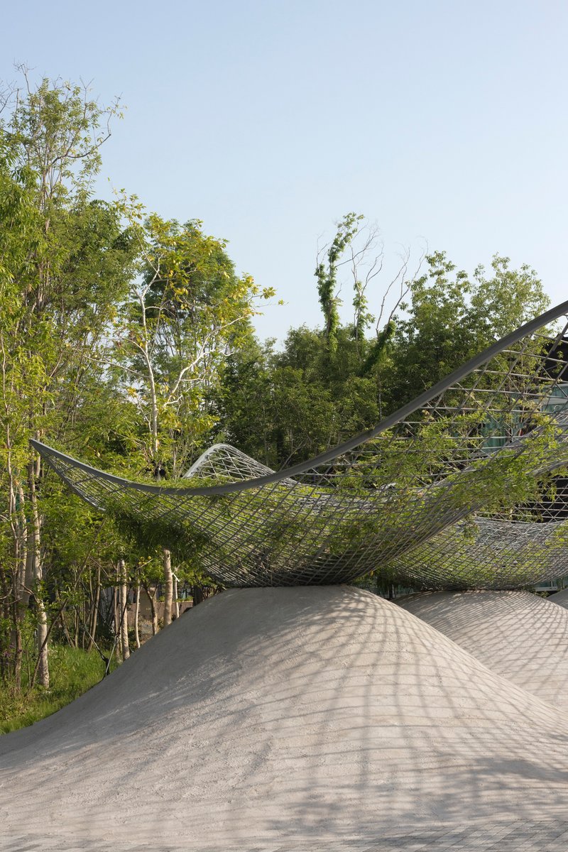 Curved concrete roof beneath a tensile steel mesh canopy suspended above leafy birch trees