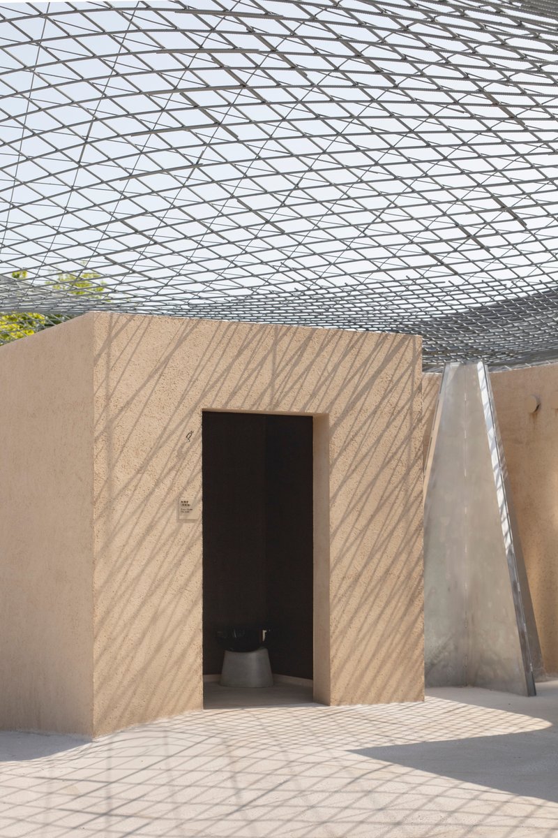Textured rammed earth doorway beneath the geometric shadow pattern cast by the overhead mesh structure
