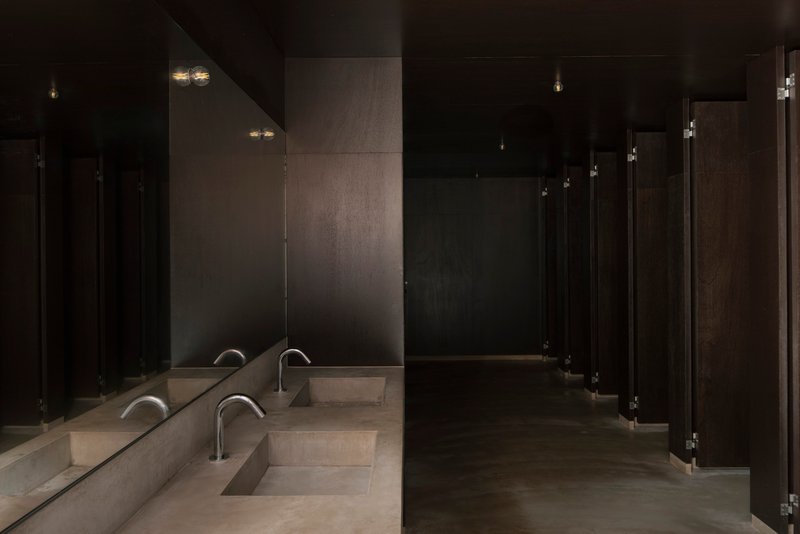 Dark restroom interior with continuous concrete trough sinks and minimalist wall-mounted faucets